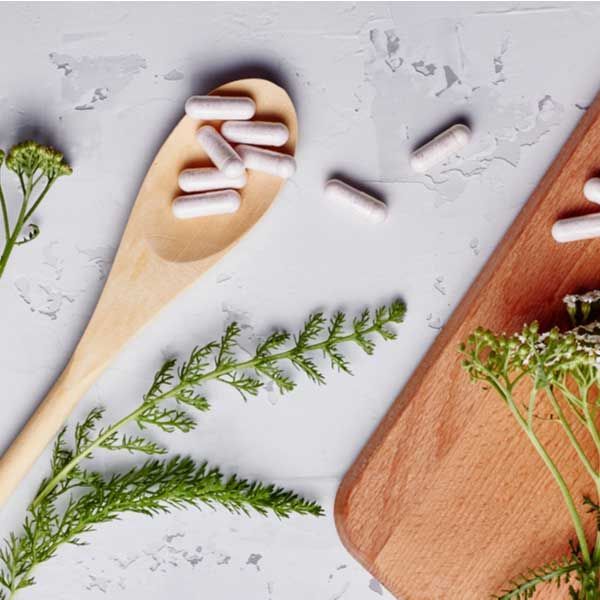 A counter, with a wooden spoon, vitamins, herbs and a cutting board on it